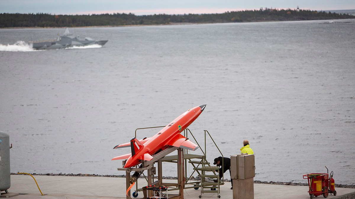 An orange coloured target drone BQM167i on the launcher, with a Visby corvette at sea in the background
